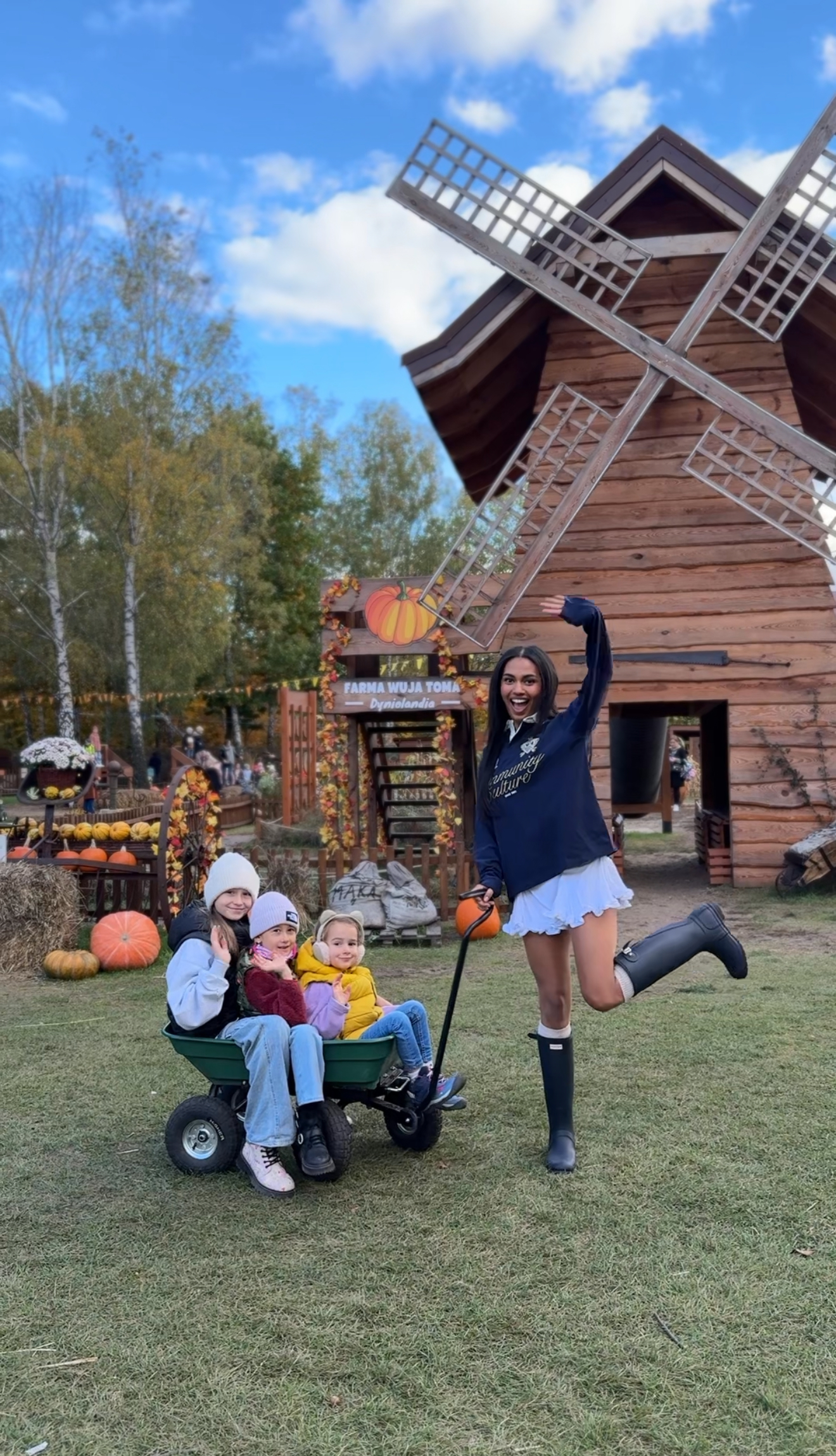 Smiling woman lifting one leg next to wagon carrying three children in front of windmill.