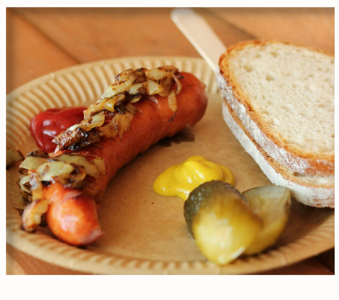 Grilled sausage topped with caramelized onions, ketchup, mustard and crusty bread on paper plate