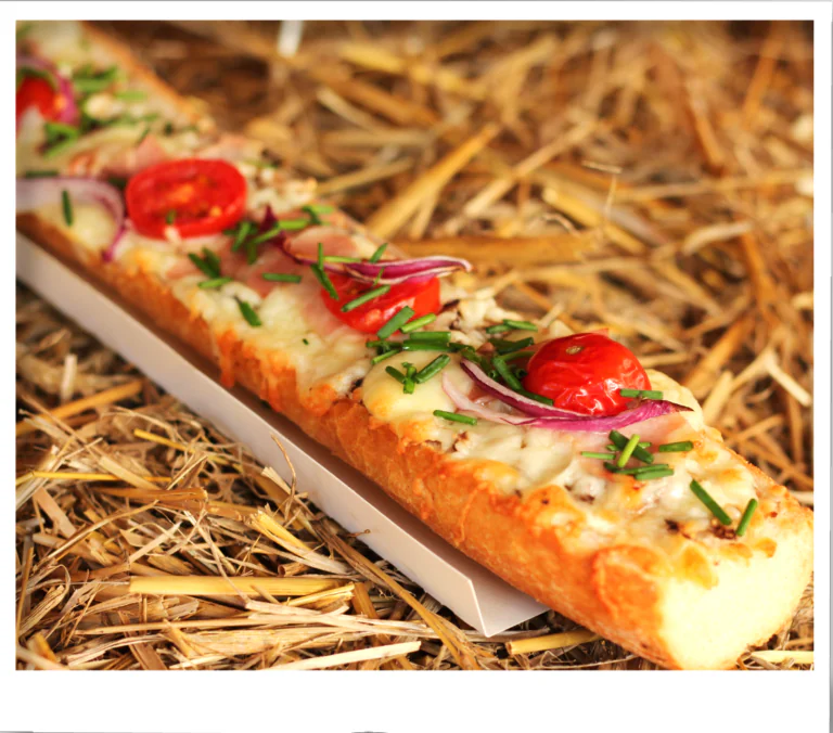 Long cheesy baguette garnished with roasted cherry tomatoes, sliced red onion, chives on straw background.