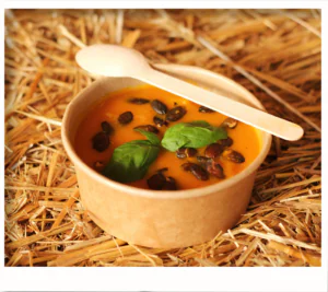 Small kraft bowl of creamy squash soup with basil and roasted seeds, wooden spoon across rim.